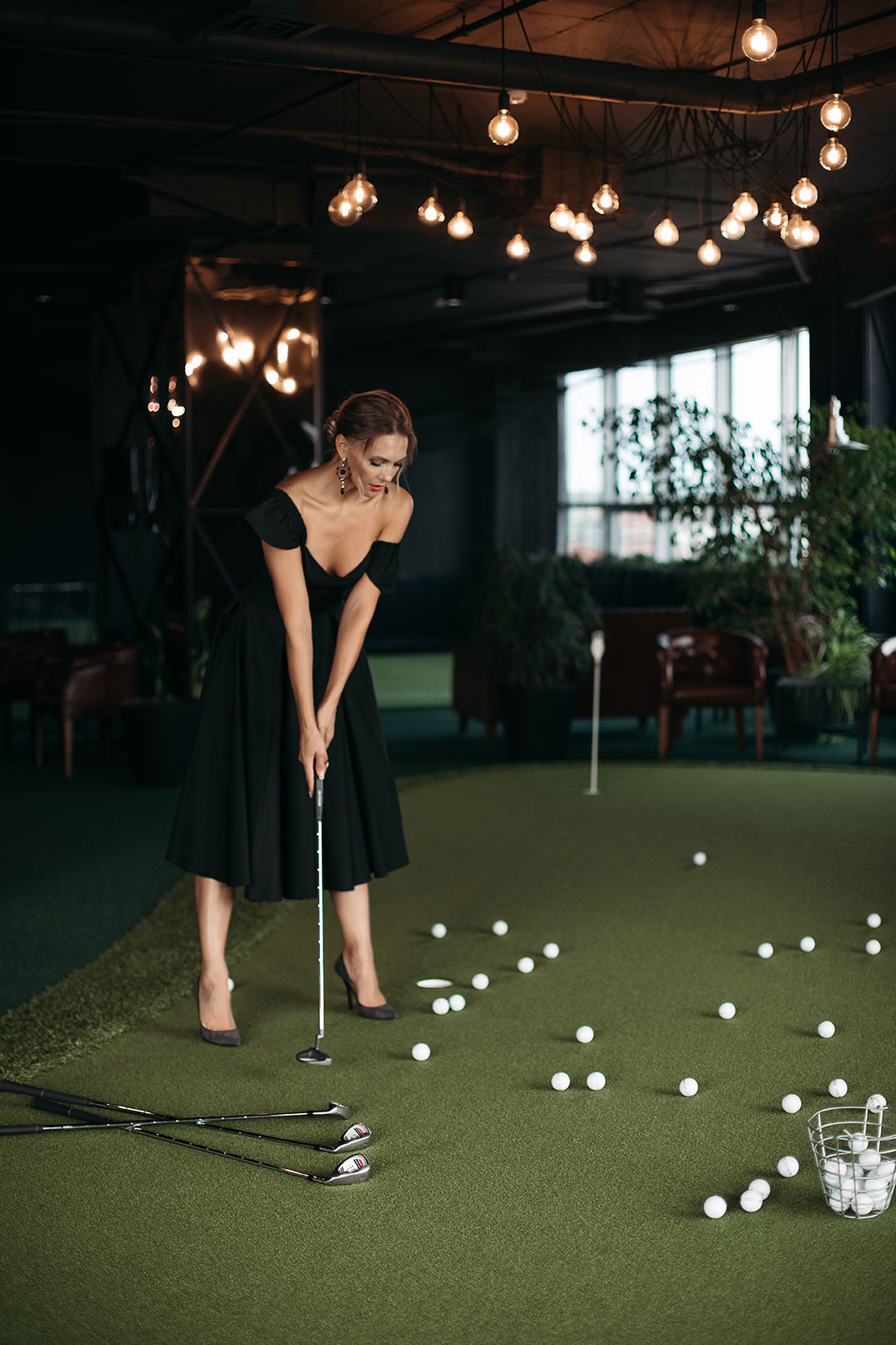 Une femme en robe de soirée et talons hauts se tient sur un tapis de gazon synthétique, bâton de golf en main, et est entourée de balles.