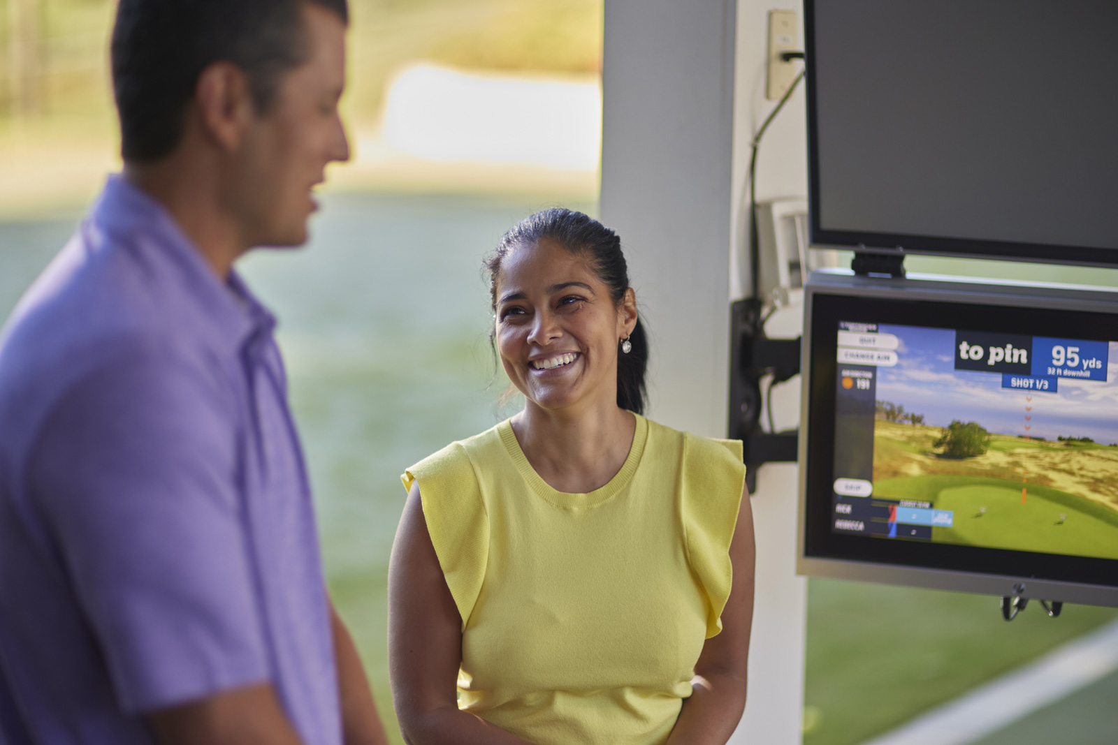 Un homme et une femme discutent en souriant. En arrière-plan, on voit un écran affichant les résultats de leurs dernières frappes de golf.