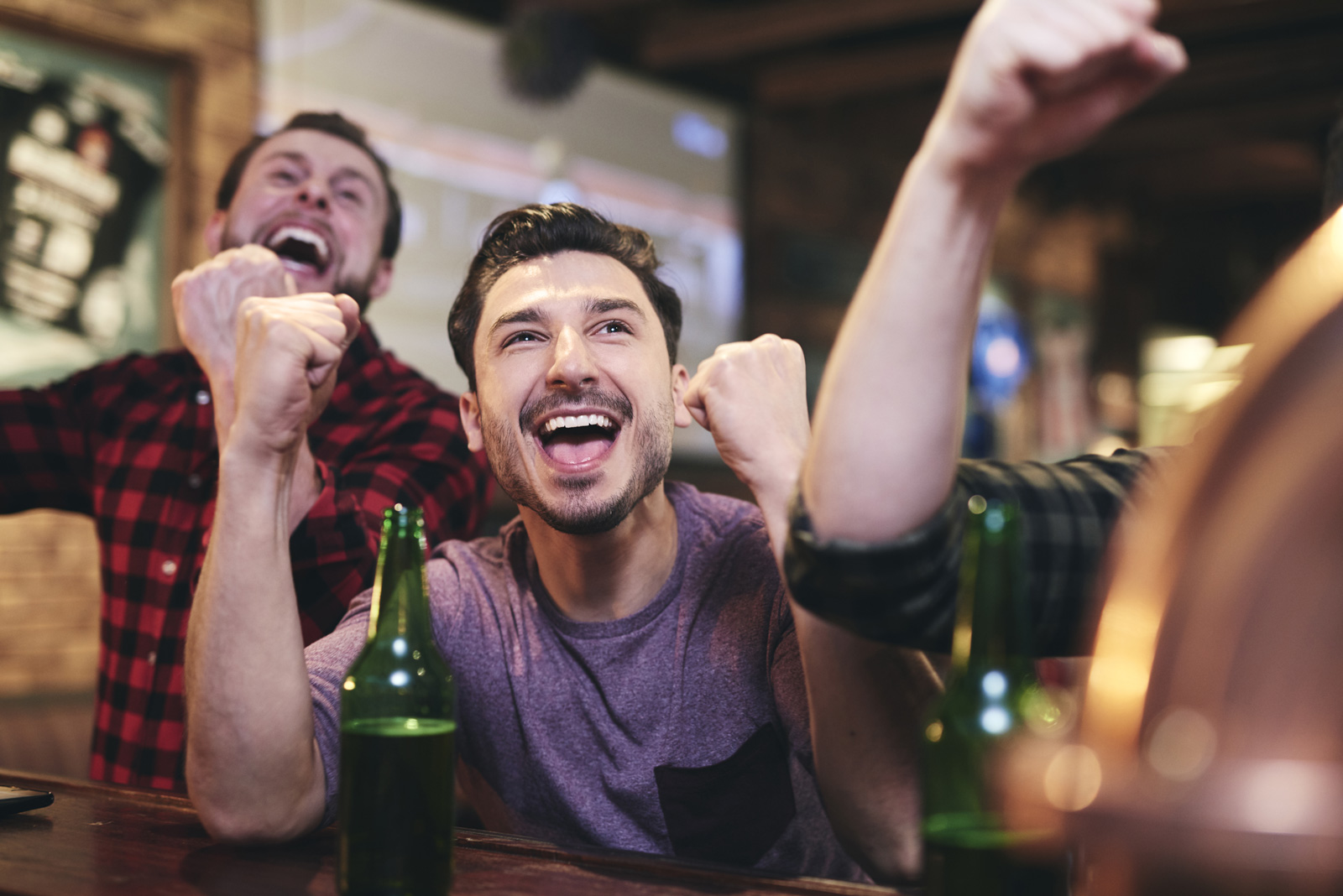 Deux hommes accoudés à un bar regardent un match de sport sur des télévisions et célèbrent une victoire dans une ambiance animée.