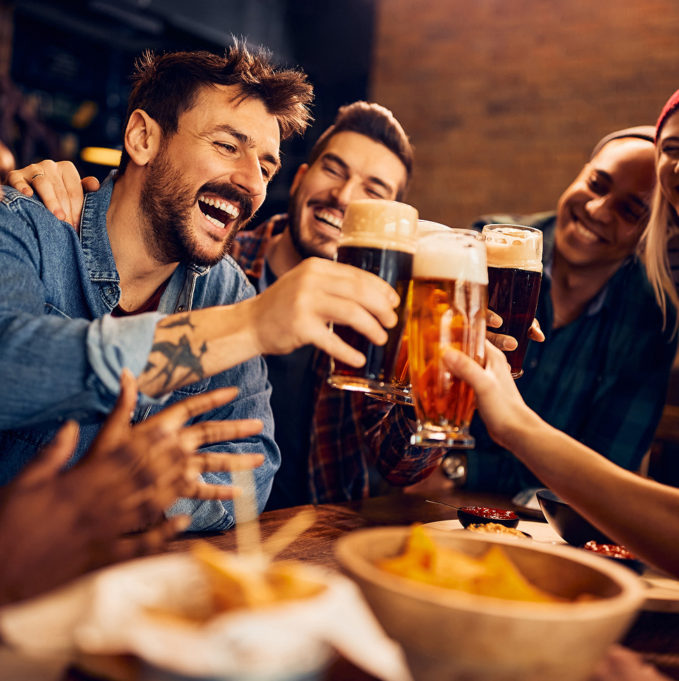 Un groupe d’amis trinquent en souriant, verres de bière à la main, dans un espace de type resto-bar.